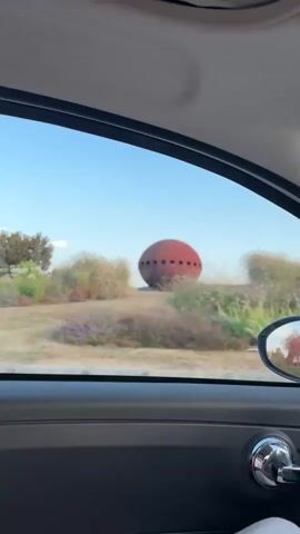 The video shows a red, oval-shaped object with dark circular markings, possibly windows, situated in a dry, grassy landscape with some bushes and a tree in the background. The object appears to be a sculpture or a decorative structure, possibly resembling a UFO or an egg. The camera is inside a vehicle, looking out the window. A person's arm and shoulder are visible in the foreground, with the person's face partially visible on the left side of the frame, looking towards the object. The sky is blue with some clouds, and the lighting suggests it is either early morning or late afternoon. The vehicle is moving, as indicated by the slight blur of the background and the motion of the scene. There is a blue and white circular sign visible on the right side of the frame in one of the shots. The word "Google" is partially visible on a sign behind the red object in one segment of the video.