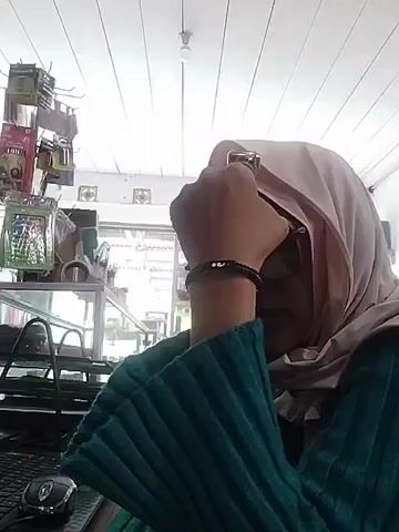 A close-up shot shows a person wearing a light-colored hijab and a teal ribbed shirt. The person's right hand is raised to their face, holding a gold-colored object near their eye. A black bracelet with white beads is visible on their wrist. The background includes shelves with various items, suggesting an indoor retail environment. The ceiling is white and appears to be made of parallel panels with a light fixture in the center.