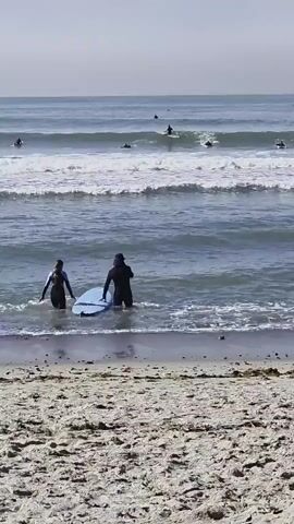 The video shows a beach scene with people surfing in the ocean and standing on the sand. In the foreground, two surfboards are lying on the sand. A person wearing a wetsuit and a blue surfboard stands in the shallow water. Further out in the ocean, several surfers are riding waves. On the beach, three people are gathered near surfboards. One person is facing away from the camera, wearing a patterned wetsuit. Another person is wearing a bikini, and the third person is wearing a baseball cap and a light blue shirt. The ocean has several rows of waves, and the sky is overcast. A seagull is visible on the sand in the middle ground.