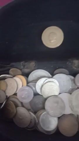 A close-up, shaky video shows a pile of coins. A single coin with a profile of a person's head is in focus at the top of the frame, against a dark background. Below it, out of focus, is a jumbled pile of various coins. The lighting is dim.
