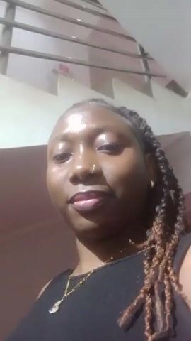 A woman is seen in a close-up, looking directly at the camera and smiling. She has dark skin, braided hair, and is wearing a black tank top with a gold necklace and a nose ring. The background consists of a pinkish-brown wall and a white structure that appears to be part of a staircase or other architectural element. The lighting is somewhat bright, causing a shine on her face. The woman says "Hi".
