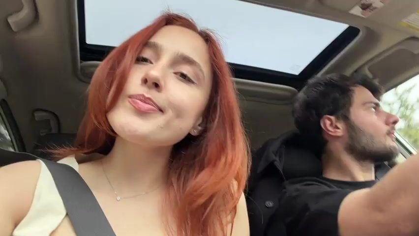 A young woman with shoulder-length auburn hair sits in the passenger seat of a car, facing the camera. She wears a seatbelt and a light-colored top, along with a delicate necklace. To her right, the driver, a young man with dark hair and a beard, is visible. Both individuals are in their early to mid-twenties. The car has a sunroof, allowing natural light into the vehicle. The woman looks at the camera and smiles, her expression calm and content. The man keeps his focus on driving.