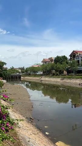 The video pans across a canal lined with buildings and trees under a clear blue sky with scattered clouds. On the left, the canal bank is made of concrete and dirt, with some debris visible. The water in the canal is murky green and reflects the sky and surrounding vegetation. Along the right bank, there is a row of houses and trees. A white floating structure with a green roof is prominently featured, supported by wooden stilts and blue buoyant barrels. A red boat with a weathered hull is docked next to this structure. The buildings in the background are varied in style and color, with some appearing to be under construction. There are no people visible in the video.
