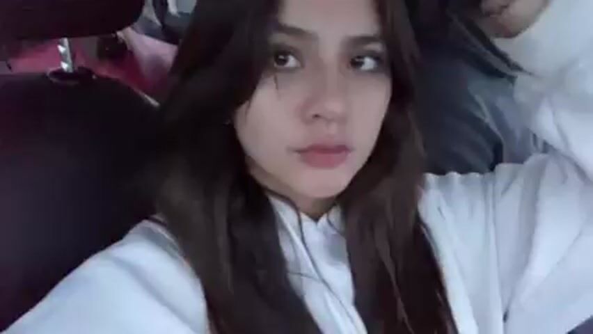 A young woman with long brown hair is in the passenger seat of a car. She is wearing a white hoodie.  A man is driving the car. The car is moving and the scenery is visible through the window. The woman smiles. There is music playing in the background.
