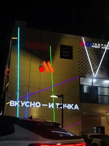A nighttime exterior view of a modern building is illuminated by colorful neon lights and building signage. The building has several levels and features large windows. A prominent sign in white lettering across the lower section of the building reads "ВКУСНО – И ТОЧКА," which translates to "Tasty - and that's it." Above this, a yellow logo with an orange circle and two curved orange shapes is visible. To the right of the logo, a sign in white lettering reads "INSPOT КИБЕР АРЕНА," indicating a cyber arena. Further up, a smaller black and white sign says "INSPOT КИБЕР СПОРТ." Geometric patterns of white neon lights form a 'V' shape on the right side of the building. Various colored neon lines, including blue, purple, and red, crisscross the building's facade. In the foreground, the rear of a white car is visible, with its taillights illuminated. Another white car is partially visible on the right side of the frame. The overall scene depicts a brightly lit commercial establishment at night.