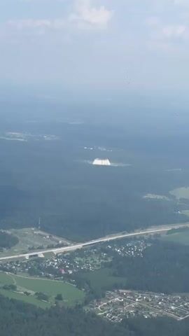 An aerial view of a landscape with dense green trees, a highway, and several small residential areas. In the distance, there is a large, white, rectangular structure that appears to be a building or a mound of some kind. The sky is hazy with a few scattered clouds. There are no people or animals visible. The overall impression is one of a rural or semi-rural environment viewed from a high vantage point.