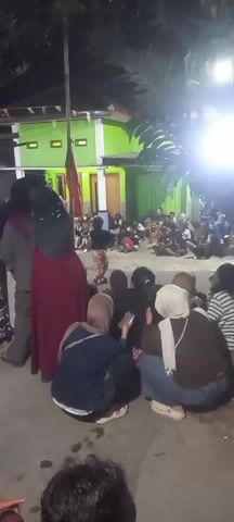 The video shows an outdoor gathering at night. In the foreground, several people are sitting or crouching, facing away from the camera. They appear to be watching an event. In the middle ground, a person dressed in traditional patterned pants and a dark top is standing and gesturing with their arms. Behind this person, a crowd of people are seated on the ground, also observing the event. To the left, a tall pole with a red flag is visible. In the background, a green building with lights is illuminated. The overall atmosphere seems to be that of a community event or performance.