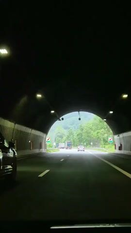 The video shows a view from inside a vehicle as it exits a dark tunnel. The tunnel has overhead lighting fixtures. Outside the tunnel, there is a multi-lane highway with a green, mountainous landscape on the right. Several other vehicles, including cars and trucks, are visible on the highway. The sky is overcast. The focus of the video is the road ahead and the scenery.