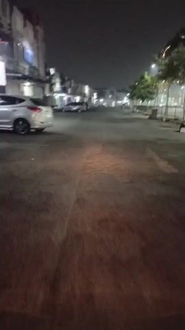 A first-person perspective video shows a street at night. Cars are parked on the left side of the street. Streetlights illuminate the road, casting a warm glow. Trees line the right side of the street, their leaves appearing as blurry green shapes against the dark sky. The camera moves forward at a consistent speed, creating a sense of motion. The overall atmosphere is calm and quiet.