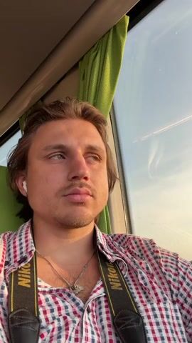 A young man with blond hair, a mustache, and earbuds is sitting inside a vehicle. He is wearing a red and white checkered shirt and a camera strap around his neck. The man looks out the window at the sky and a rural landscape. The vehicle appears to be in motion, indicated by the blurry movement of the scenery outside the window. The sun is setting, casting a warm glow on the scene.