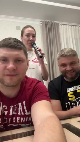 The video shows three people singing karaoke. A man wearing a red shirt holds a camera, filming himself and the others. Behind him, a woman holding a microphone sings, and a man in a black shirt sits beside the cameraman, initially looking displeased but then smiling slightly. The scene appears to be indoors, possibly in a home, with a table and curtains visible in the background. The lighting is bright.