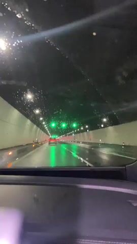 The video shows a first-person view from inside a car driving through a tunnel. The tunnel is illuminated by a series of green lights overhead, which reflect off the wet road surface. Water droplets are visible on the windshield, suggesting it is raining outside or the tunnel is humid. The car is in motion, and other vehicles can be seen ahead, their taillights glowing red. The tunnel walls are white and appear to be made of concrete. The ceiling of the tunnel is dark and speckled with lights.