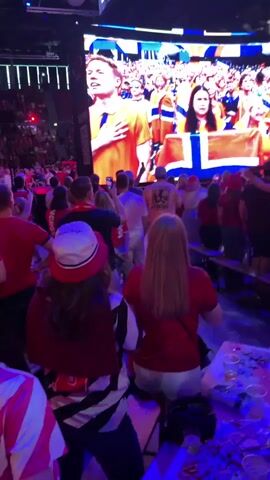 This media file captures a lively scene from a sporting event or concert, likely in Oslo, Norway, given the presence of Norwegian flags. The primary subjects are a large crowd of people, many wearing red, and a massive screen displaying images of athletes or performers.

The scene is set within an indoor arena or stadium, illuminated by vibrant stage lighting that casts a purple hue across the crowd. The screen dominates the background, showing individuals in red attire, some holding a Norwegian flag, suggesting strong national pride or support for a team. The crowd is densely packed, with people standing and facing the screen, indicating they are actively engaged in watching the event.

In the foreground, several individuals are visible, their backs mostly turned to the camera. They appear to be part of the audience, some holding drinks. The atmosphere seems energetic and celebratory, with the overall mood appearing to be one of excitement and communal enjoyment. The time of day is not explicitly discernible, but the indoor setting and artificial lighting suggest it could be evening or night.

There is no discernible text within the media file itself. The focus is on the collective experience of the audience and the spectacle unfolding on the large screen.