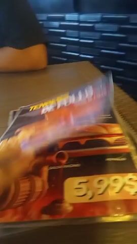 The video shows a person's hands flipping through a set of laminated menus on a table. The menus are brightly colored and feature images of food items like burgers, chicken tenders, and pizza, along with prices in dollars. The background includes a textured wall and a glimpse of another person sitting at the table. The menus are bound with a spiral.
