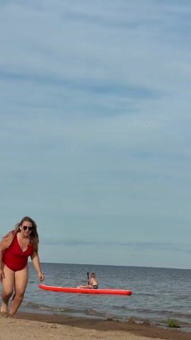A full shot video shows a woman running along a sandy beach on a clear, sunny day. She is wearing a red one-piece swimsuit and sunglasses. In the background, a young boy sits on a red paddleboard in the ocean, facing away from the camera. The sky is blue with scattered white clouds. The water is calm, with gentle waves lapping at the shore. The woman appears joyful and energetic as she runs towards the camera. The scene suggests a casual summer day at the beach, with activities like paddleboarding and running for recreation.