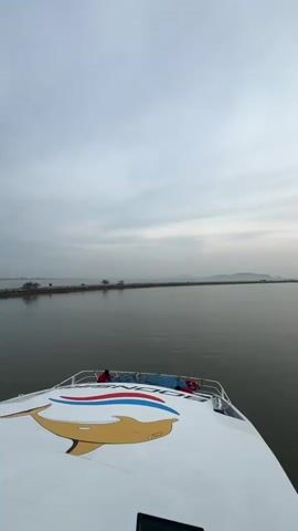 The video is captured from the deck of a boat, looking towards the horizon. The boat's deck is white with a large drawing of a dolphin. Two people are visible in red shirts on the deck above. The water is calm with a gray hue, reflecting the overcast sky. In the distance, a land mass is visible, appearing as a long, narrow strip with some trees. To the right, there's another land mass with a small building or structure, possibly a lighthouse. As the video progresses, the sun begins to peek through the clouds, casting a reflection on the water.