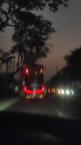 The video shows a blurry nighttime view from inside a vehicle, looking forward at a road. A large bus with red taillights is visible ahead, appearing to be in motion. Several other vehicles with lights are also seen on the road, some appearing to be on the right side of the bus and some in the distance. Trees are visible on the left side of the road, and power lines can be seen above them. The sky is dark, suggesting dusk or night. The overall impression is of traveling on a road at night, possibly in a rural or semi-rural area.