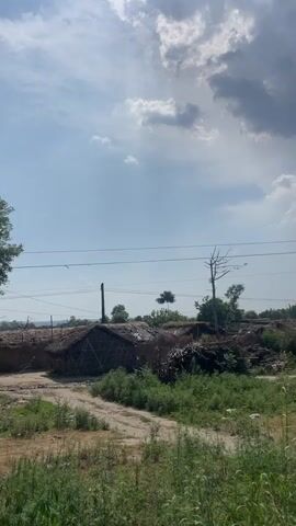 The video shows a landscape with a few simple huts and a dirt path. The huts appear to be made of natural materials like straw or reeds. A pile of branches and vegetation sits near one of the huts. Green grass and weeds cover the ground. Power lines stretch across the sky, which is partly cloudy with patches of blue. A few trees are visible, one of which is bare. The camera pans slightly to the right, revealing more of the landscape.
