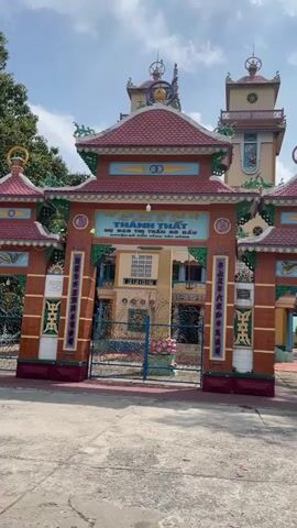 The video shows a temple gate with a red tile roof and a brick facade. The gate is closed and leads to a building with a tower in the background. The sky is blue with some white clouds.