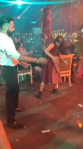A crowd of people are dancing on a stage, and confetti is falling from the sky. Two women are dancing in the center, one on the ground and one standing on the other's shoulders. The woman on the ground is wearing a dark dress with a plaid skirt. The woman on her shoulders is wearing a dark dress. A man in a white shirt and dark pants is standing to the side, watching. There are chairs and tables scattered around the stage, and people are sitting and standing around them. There are colored lights shining on the scene.