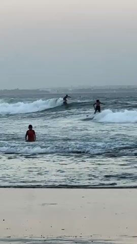 This video shows a beach scene with several people in the ocean. In the foreground, a person in an orange shirt stands in shallow water. Further out, there are multiple surfers riding waves. One surfer is prominently positioned in the middle of the frame, riding a wave. Another surfer is visible to the right. The ocean has choppy waves, and the sky is overcast. The beach in the foreground appears sandy, with some debris, possibly plastic, visible on the wet sand. The overall atmosphere is of a casual day at the beach with water activities.