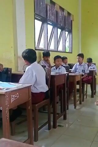 The video depicts a classroom setting with several young students, likely elementary or middle school age, seated at wooden desks. Most students are wearing white shirts and red pants or skirts, suggesting a school uniform. The classroom has yellow walls and a large window with wooden blinds. There are papers and books on the desks. In the foreground, a student wearing a white hijab is reading a book. To her right, another student in a white hijab is resting their head on their arm. Further back, several other students are visible, some looking towards the front of the classroom or at their books. One student is standing at a desk in the background, appearing to handle some papers. The overall impression is of a typical classroom environment during a lesson or study period.