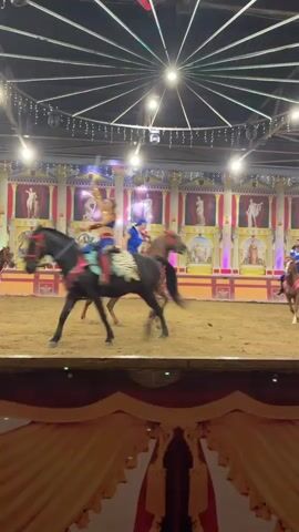 The video shows a horse show in an arena. Three horses are visible, two of which are ridden by individuals in costumes. The horses are moving at a trot or canter. The arena appears to be indoors, with a dirt floor and a decorated backdrop featuring statues and paintings. Lights are suspended from the ceiling, and a fabric curtain is visible in the foreground. The riders are wearing brightly colored outfits, and one rider appears to be holding a lance or flag. The overall scene suggests a performance or exhibition.