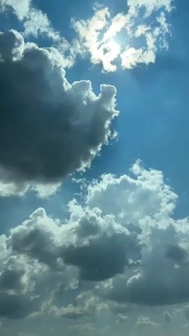 The media file displays a dynamic sky with dramatic clouds and the sun peeking through. The scene is set outdoors, with a clear blue sky dominating the upper portion. Puffy, white clouds are interspersed with darker, more ominous-looking clouds, suggesting a mix of weather conditions. The sun is bright and partially obscured by clouds, casting rays of light that create a sense of depth and drama in the sky. Some of the clouds appear to be cumulus or cumulonimbus clouds, indicating potential for rain or stormy weather.

The time of day appears to be daytime, given the brightness of the sun. The location cues suggest an outdoor setting, possibly a rural or open area, as there are no urban elements visible. The overall mood conveyed by the image is one of awe and natural beauty, with the interplay of light and shadow adding a sense of majesty. There is no visible text in the media file. The weather appears to be partly cloudy with bright sunshine.