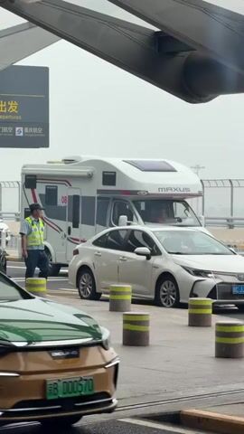 The video shows a traffic scene from the perspective of a stationary green and gold car. In the foreground, the hood and windshield of the green and gold car are visible. Behind it, a white sedan is driving. Further in the background, a white RV with red accents and a graphic on the back is visible, along with other vehicles, including a white van with flashing lights and a dark-colored SUV. A person in a yellow vest and a dark helmet is standing near the RV. The scene appears to be at an outdoor location, possibly an airport or a toll booth, with overhead structures and railings visible. The sky is overcast.