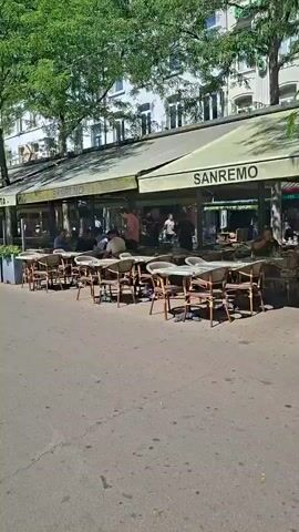The video shows an outdoor seating area of several restaurants or cafes. Tables and chairs are set up on a paved walkway, with awnings providing shade. Some of the tables have umbrellas. People are seated at some of the tables, eating and talking. Trees line the walkway, and buildings are visible in the background. The overall impression is of a pleasant, sunny day in a busy urban area.