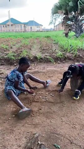 In a daytime outdoor scene, two young girls are engaged in play on a dirt ground in Riyom, Nigeria. The sky appears overcast, providing diffused light. One girl, wearing a blue and white patterned dress and sandals, sits on the ground with her legs extended. She is focused on a pattern drawn in the dirt, surrounded by small stones or shells that are part of their game. The other girl, dressed in black pants and a black top with neon green accents on her shoes, crouches or stands beside a wooden log, also interacting with the stones on the ground. She appears to be knocking over some of the stones. In the background, a cultivated patch of ground with green plants is visible, along with several residential buildings and a tall palm tree under a cloudy sky. The girls seem absorbed in their game.