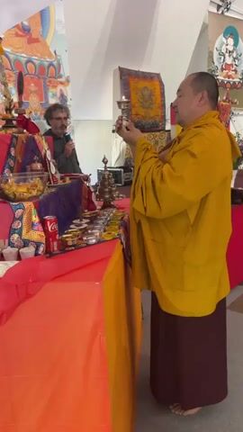 The video shows a monk in a yellow robe holding a lit lamp and reciting in a room. He is holding the lamp with two hands close to his chest. He has a shaved head, and he is wearing maroon pants under his robe. He appears to be standing near a table covered with colorful fabrics, offerings, and a can of Coca-Cola. Another man with a microphone stands behind the table. A second monk stands in the background. The room seems to be a part of a temple or religious center, with religious paintings visible on the walls. The scene appears to be a religious ceremony or prayer ritual.