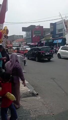 A street scene with several people standing on the sidewalk. There are cars and motorbikes on the road. Balloons are visible on the left side of the frame, attached to a pole. A sign that says "OPPO" is visible on a building in the background. The sky is overcast.