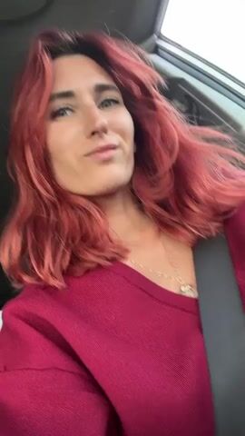 A woman with red hair, wearing a maroon sweater and a seatbelt, smiles at the camera from inside a car. The camera is positioned at eye level and slightly off-center. Light is coming from the car window.