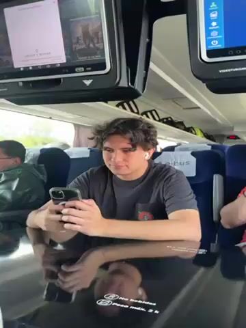 A bus passenger is seen sitting at a dark, reflective table in a bus. He has dark curly hair and is wearing a black t-shirt. He is looking at his cell phone, which he holds with both hands. He wears wireless earbuds. The table reflects his face. Other passengers can be seen. Two television monitors hang above the tables. The monitors show various options, including movies, to the passengers. Text is visible on the table, below his reflection. "socibus" is printed on the seats. The window is partially visible.