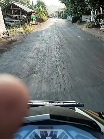 The video shows a first-person perspective of someone riding a motorcycle or scooter. The rider's thumb is visible in the foreground, partially obscuring the view. The dashboard of the vehicle is also visible, with a speedometer. The road ahead appears to be paved and somewhat rough, with cracks visible in places. The environment is rural or suburban, with small buildings and vegetation lining the road. In the distance, other vehicles, including a truck and another motorcycle, are visible. The sun is shining, casting long shadows. The overall impression is of a daytime journey through a residential or semi-rural area.