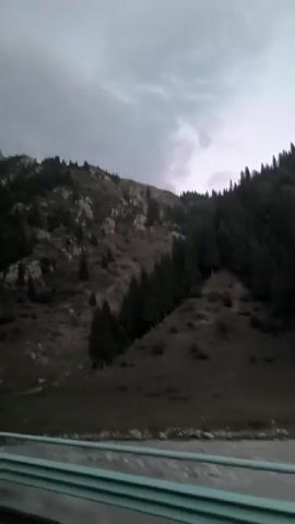 The video shows a mountainous landscape from the perspective of a moving vehicle. The sky is overcast with dark clouds. The mountains are rocky and covered with sparse vegetation, including some evergreen trees. A road with a blue and white barrier is visible in the foreground. The lighting suggests it is either early morning or late evening, or a heavily overcast day.