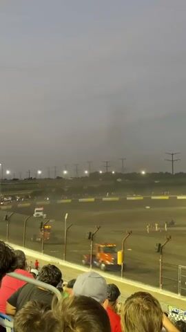 The video shows a dirt racetrack from the perspective of spectators in a grandstand. Several tow trucks are on the track, with one appearing to tow a vehicle. A few people are visible on the track near the tow trucks. The sky is overcast, and artificial lights are illuminating the track. The spectators in the foreground are mostly out of focus, with some heads and shoulders visible. The overall atmosphere suggests a racing event that may have been interrupted or is in a downtime.