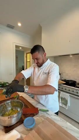A man in a white chef's coat with the word "CHEFB" visible on the left breast is standing in a kitchen. He is wearing black gloves and a watch on his left wrist. He is holding a lime in his gloved hands and squeezing it over a metal bowl filled with a green mixture, presumably guacamole. The bowl is on a wooden cutting board. To the left of the bowl are some chopped green herbs. To the right of the bowl are various kitchen items including a blue-lidded container, a wooden cutting board, and a white towel with a palm leaf design. The kitchen has white countertops and white cabinetry, with a stove visible in the background. The lighting suggests it is daytime.