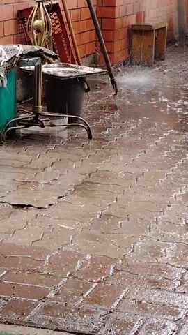 The video shows a courtyard paved with interlocking bricks. A person is power washing the pavement with a high-pressure washer, creating a spray of water. The water is making the bricks wet and shiny, and some of it is splashing up. There are various objects in the background, including a brick wall, a wooden bench, a bucket, and some gardening tools leaning against the wall. The lighting suggests it could be daytime, and the presence of water suggests recent or ongoing rain, or the activity of washing. The scene appears to be an outdoor residential or service area.