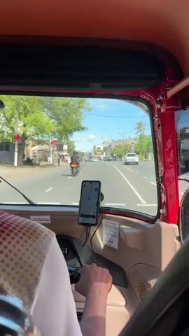 The video shows a first-person perspective from inside a red tuk-tuk, looking out through the windshield onto a sunny road in Delkanda, Sri Lanka. The driver, wearing a white shirt with red and yellow polka dots and a dark cap, is seen from behind, with their hands on the tuk-tuk's handlebar. A smartphone displaying a navigation map is mounted on the dashboard. The road ahead has light traffic, including motorcycles and cars, with buildings, shops, and trees lining both sides. Visible text includes "08 A 700 800" on a sticker and unreadable text on a small white card attached to the dashboard. The weather is clear and bright, indicating daytime.