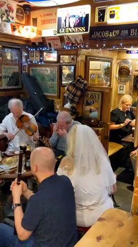 The video shows a lively scene inside a pub. A group of people are gathered around a table, enjoying live music. On the left, a man in a white shirt plays the violin. Next to him, a man in a blue shirt plays a harmonica. In the center, a man in a blue vest and collared shirt strums an acoustic guitar. To his left, another man plays a banjo. In the foreground, a man with a dark shirt and blue jeans sits with his back to the camera, holding what appears to be a wooden flute. A woman in a white top and sparkly skirt sits across from the banjo player, holding and playing a bodhrán, an Irish drum. Several glasses of beer are visible on the table. The pub is decorated with numerous framed pictures, signs, and string lights, giving it a cozy and traditional atmosphere. The lighting is warm and somewhat dim, typical of a pub setting. The overall impression is of a friendly gathering with music and good cheer.
