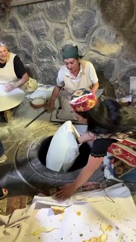 A woman wearing a black long-sleeved shirt with a red and beige patterned apron and a matching hat, kneels on the floor, leaning over a circular opening in the ground. She is placing a white, flattened dough onto the inner wall of a traditional clay oven (tandoor or tonir). Behind her, an older woman with a green head covering and a beige apron is seated on the floor, smiling and watching. To the left, another older woman with grey hair and a beige apron is seated at a low table, working with dough, while someone films her on a mobile phone. The floor is covered with white cloths, and there are pieces of flatbread on them. The background features a stone wall.