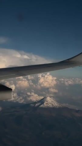 A view from an airplane shows a wing passing over the clouds and a snow-capped mountain. The sky is blue, and the clouds are a mix of white and grey. The mountain is large and prominent, with a dark, rocky base. The airplane wing is white with small lights on it. The scene is mostly still, but the airplane is moving forward. A voice is heard, but indiscernible.