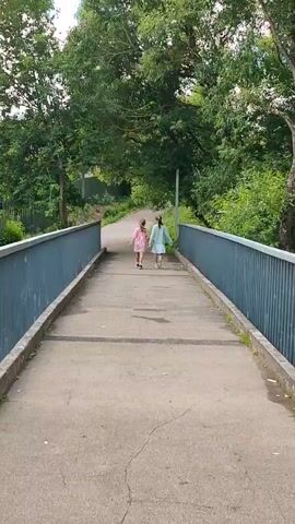 Two young girls walk away from the camera on a concrete pathway. The pathway is bordered by metal railings on both sides, and is flanked by lush green trees. The girls are walking towards a bend in the pathway, which leads into a wooded area. The girl on the left is wearing a pink dress, and the girl on the right is wearing a light blue dress. The pathway appears to be a bridge over a body of water, which can be seen between the trees on the right side of the video. There are reflections of trees on the surface of the water.