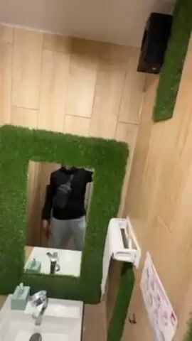 Here is a description of the video:

The video shows a bathroom interior. The walls are paneled with light wood-grain.  A mirror framed with artificial turf hangs above a white sink. A man takes a selfie in the mirror, wearing a black puffer jacket, gray pants, and a small black shoulder bag.  He is centered in the mirror's reflection, holding his phone up to take the picture.  To the right of the mirror, a matching strip of artificial turf runs down the wall.  A toilet paper dispenser and a graphic, possibly a foot diagram, are visible on the wall as well.  A black speaker is mounted in the upper right corner. The lighting in the room is bright.  Music plays in the background.