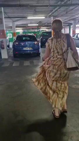 A woman with blonde hair, wearing a tan and brown patterned dress and carrying a light-colored purse over her right shoulder, walks away from the camera in a parking garage. She is walking on a concrete floor marked with white pedestrian crosswalk lines. The parking garage has a concrete ceiling with exposed pipes and fluorescent lighting. Several cars are parked in the garage, including a blue hatchback on the left, a dark SUV in the middle, and a silver sedan on the right. The woman's dress is flowing around her legs as she walks.