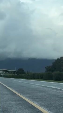 The video shows a road alongside some bushes and trees with a view of mountains partially obscured by clouds. The road is wet, likely from recent rain. The sky is overcast, with the clouds appearing gray and heavy. The mountains in the background are dark and imposing, but the low clouds shroud their peaks.