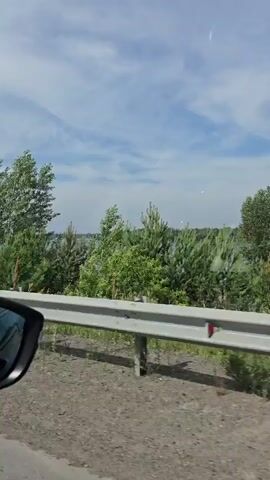 The video shows a view from a car window driving alongside a wide river. The river is calm and reflects the blue sky with scattered white clouds. Lush green trees and bushes line the riverbank, with some houses visible in the distance. A silver guardrail runs along the side of the road in the foreground. The car appears to be moving at a moderate speed, as indicated by the slight blur of the passing scenery. The overall atmosphere is peaceful and scenic, suggesting a drive through a rural or suburban area on a pleasant day.