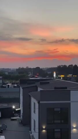 A time-lapse video shows a sunset over a modern architectural complex. The sky is ablaze with vibrant hues of orange, pink, and red, transitioning into a soft pastel at the top. Below the sky, a dark silhouette of trees and hills forms a distant horizon. In the midground, several contemporary buildings with dark gray roofs and white and dark gray facades are visible. Some buildings have external lighting fixtures that are illuminated. Cars are parked in front of some of the structures. The video captures the dynamic changes in the sky's colors as the sun descends.